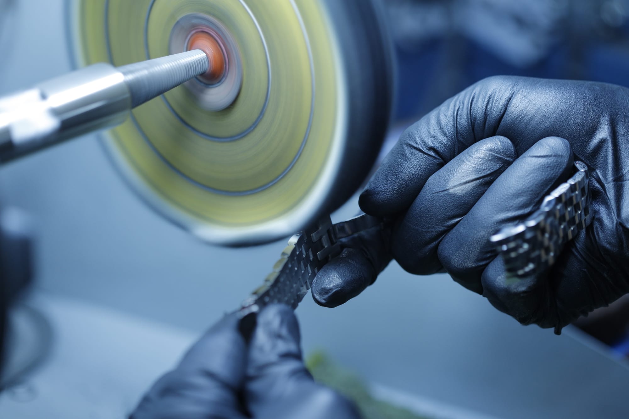 An image of a polisher's hands in black gloves holding a steel bracelet being rubbed against a buff to give a high-polish luster to the item.