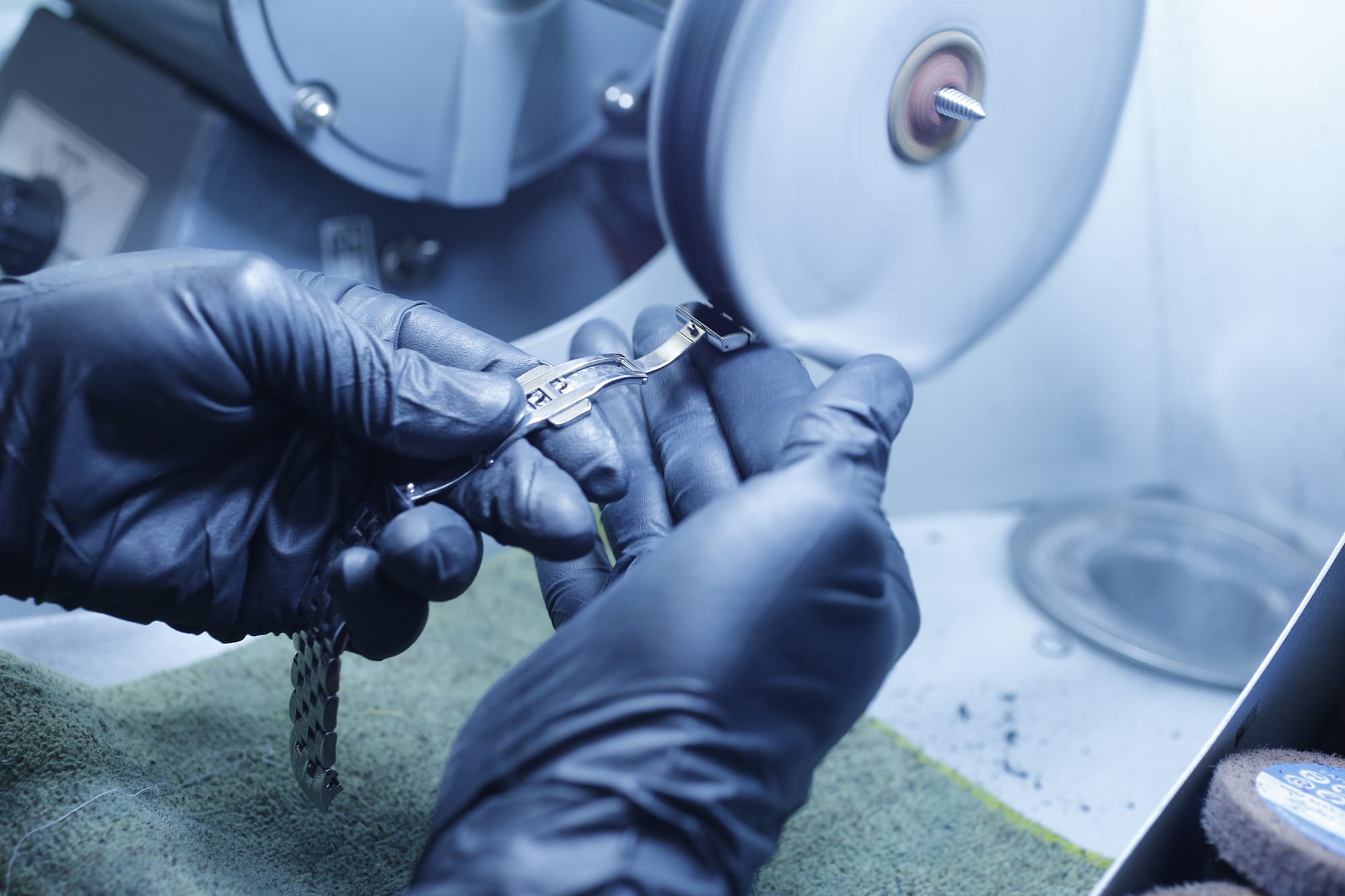 An image of a polisher's hands in black gloves holding a steel bracelet being rubbed against a buff to give a high-polish luster to the item on the top portion of the band.